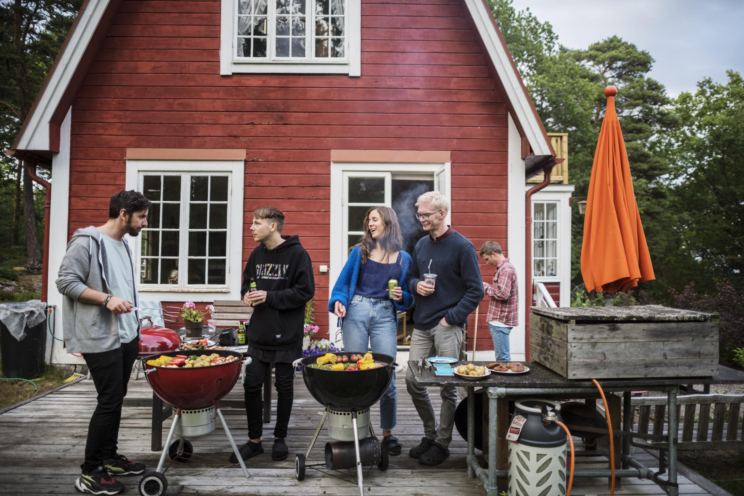 A group of people on a terrace are drinking and preparing food on two outdoor grills.